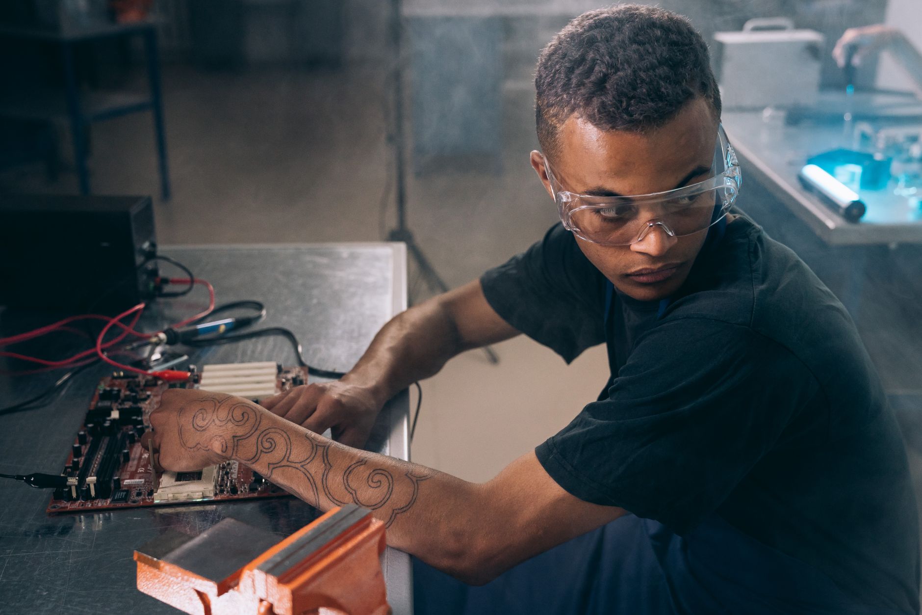 Young technician with tattoos and goggles working on electronics in a workshop setting.