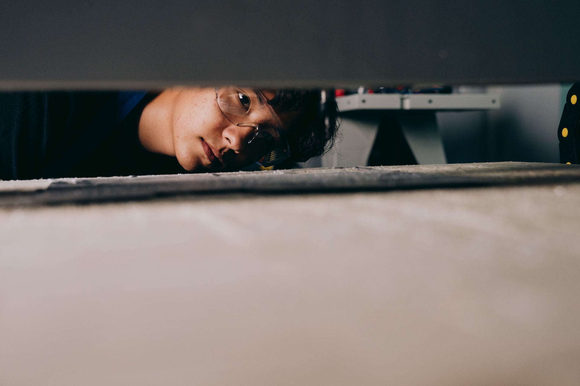 Asian man with glasses peeking underneath a wall in an indoor setting.