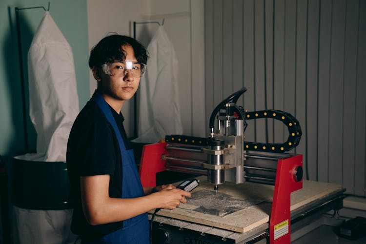 A Young Man In An Apron And Safety Glasses Standing By A CNC Machine