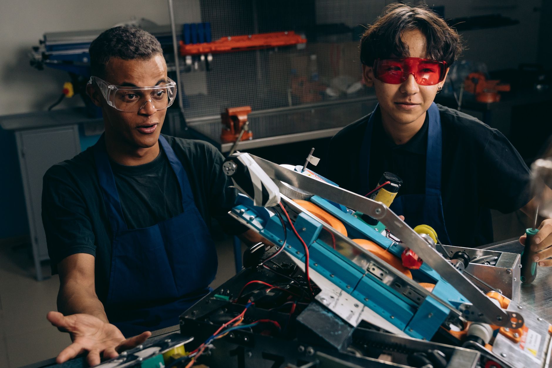 Two men wearing goggles and aprons collaborating on a robotic project in a workshop.