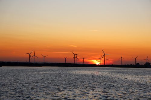 Free A scenic view of wind turbines silhouetted against a dramatic sunset sky over calm waters. Stock Photo