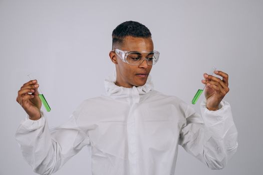 Scientist in protective gear and safety glasses analyzing green liquid in lab test tubes indoors.