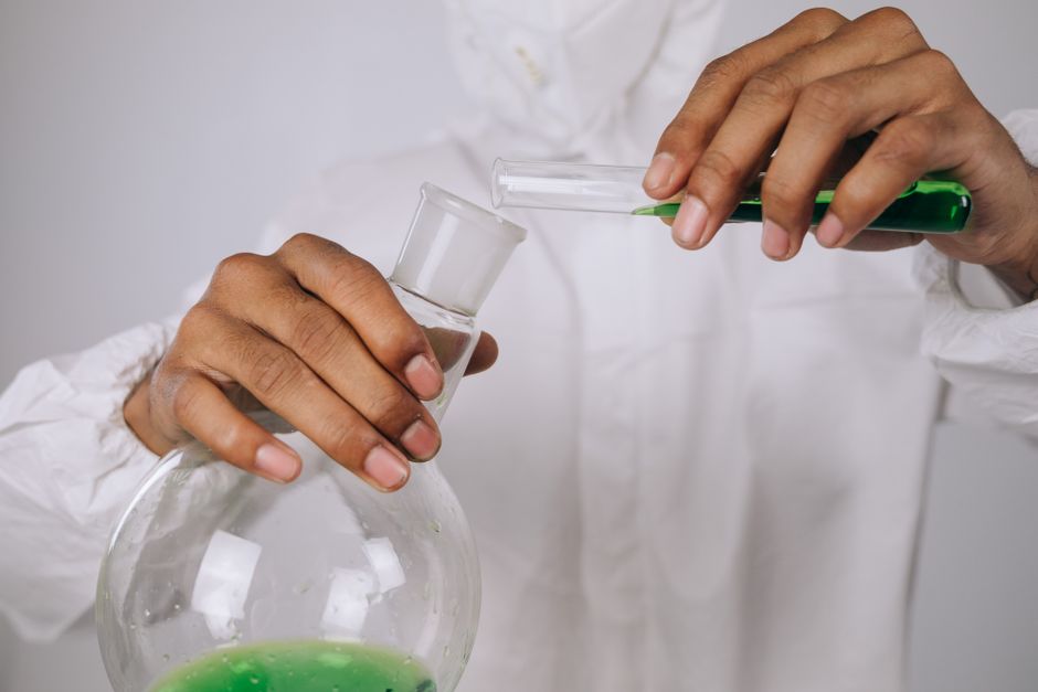Close-up of a scientist pouring green liquid between glassware in a lab.