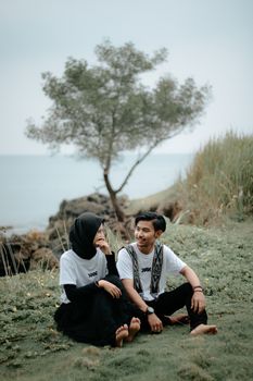 A young couple sits on a grassy waterfront, enjoying a peaceful moment outdoors. - Fotografer Sidoarjo