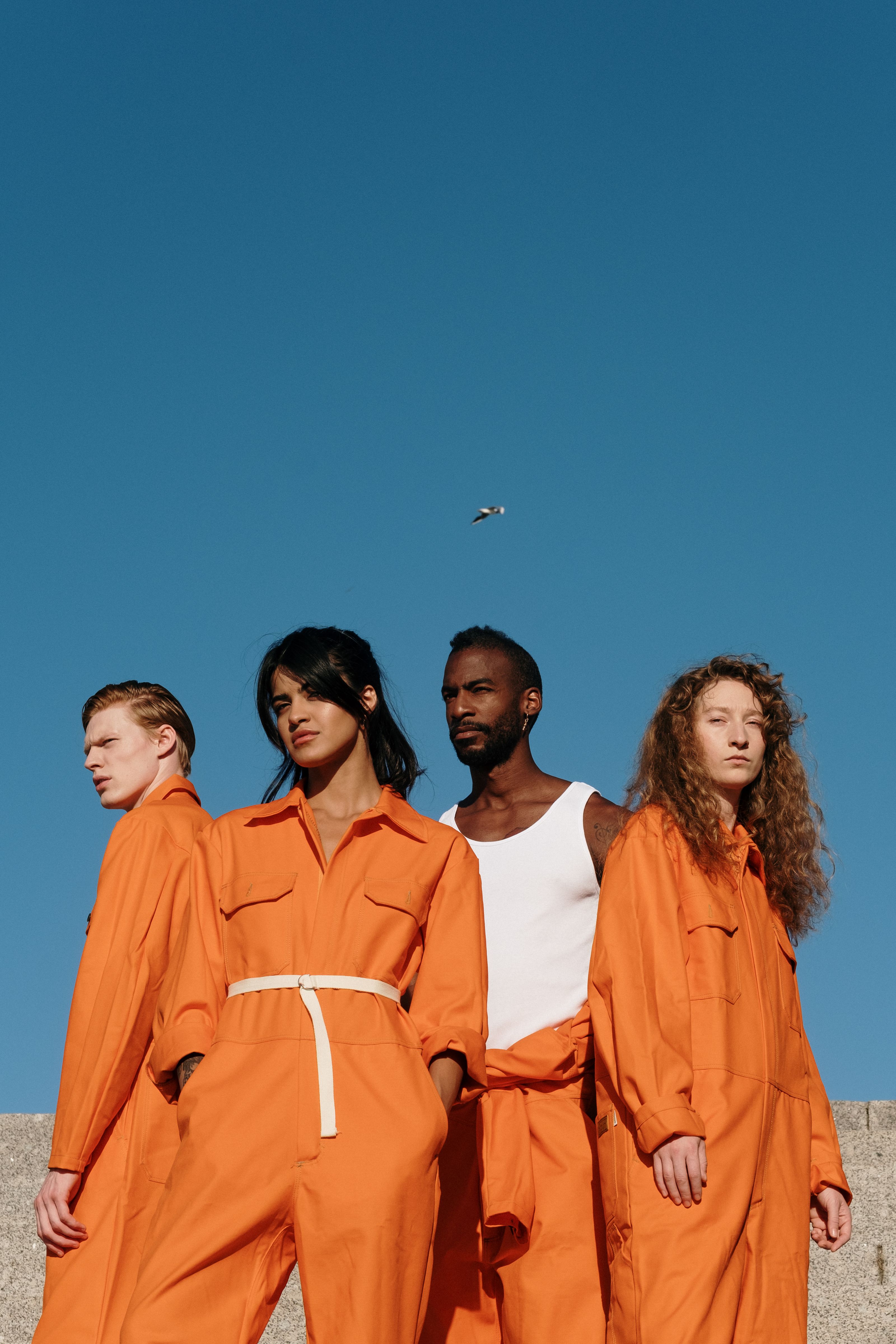Um grupo diverso de adultos em macacões laranja em pé ao ar livre contra um céu azul claro.