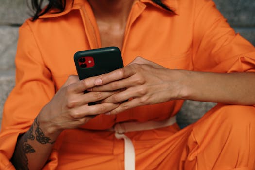 A person in an orange outfit using a smartphone, showcasing a close-up of hands and tattoo.