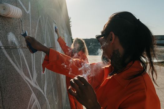 Inmates in orange uniforms painting over graffiti on a wall as part of community service outdoors.