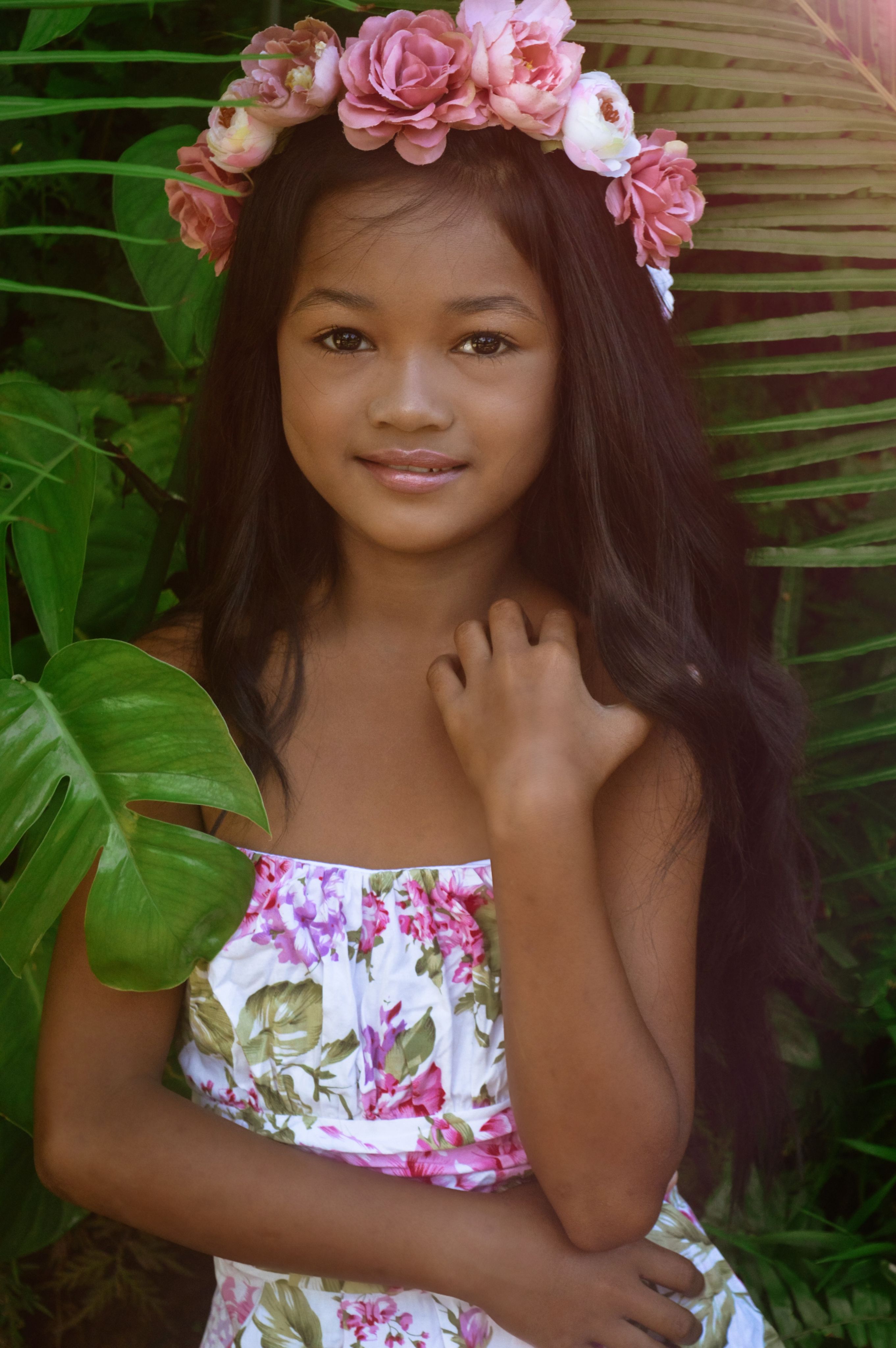 A smiling girl wearing a floral dress and flower crown poses amidst lush greenery.