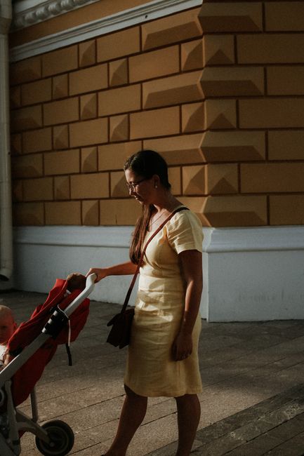 A woman in a yellow dress walks with a stroller in natural sunlight against a textured wall.
