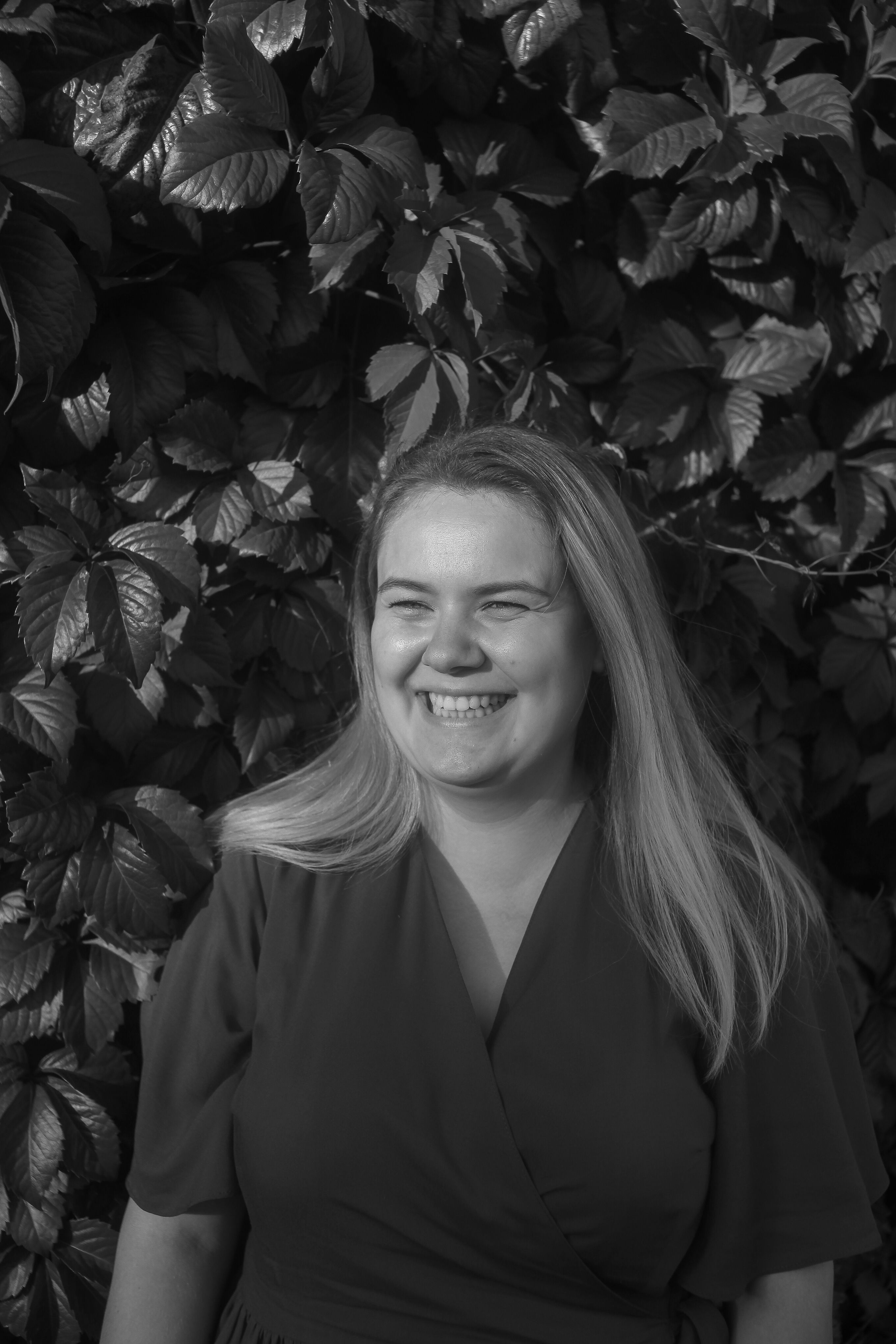 A joyful woman stands in front of lush leaves, captured in black and white.