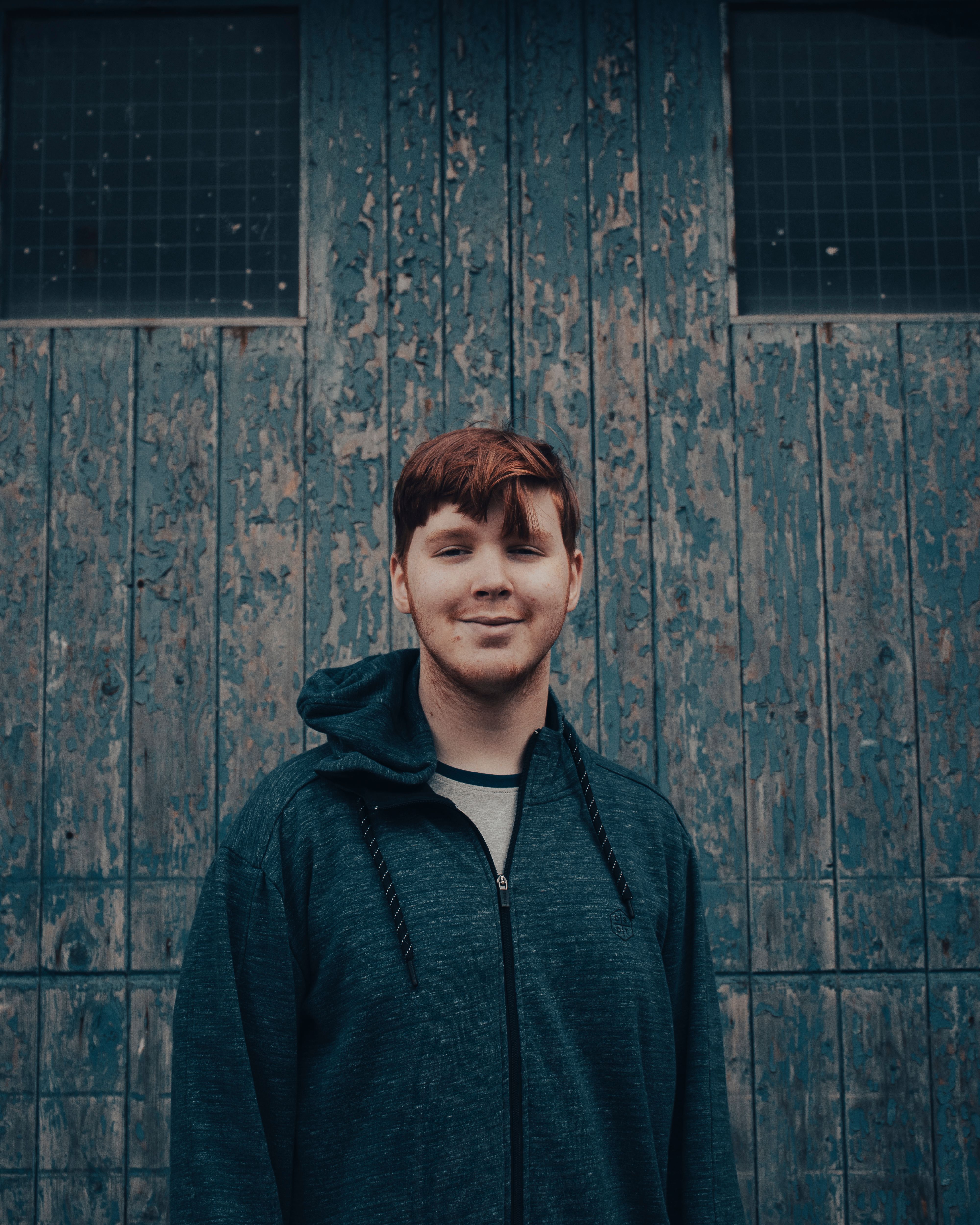 Young man in a hoodie smiling against a rustic wooden backdrop.