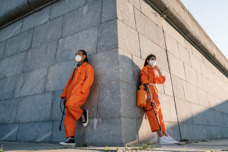 Two people in orange jumpsuits and masks leaning on a city wall.