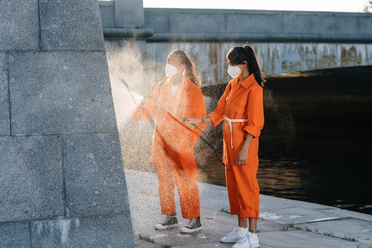 A Man And A Woman In Orange Jumpsuit Wearing Face Masks Spraying Water On A Wall