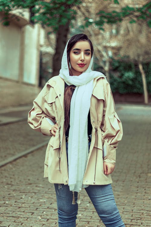 Woman in Beige Coat and White Scarf Standing on the Street while Smiling at the Camera