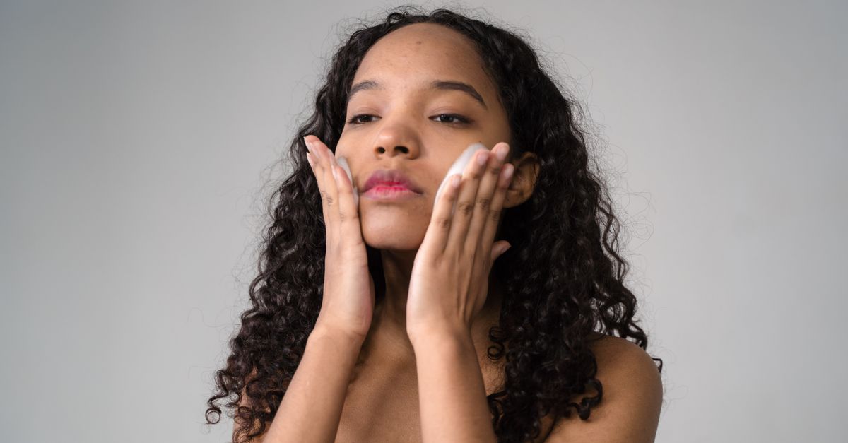 A young woman with curly hair gently applies skincare cream to her face.