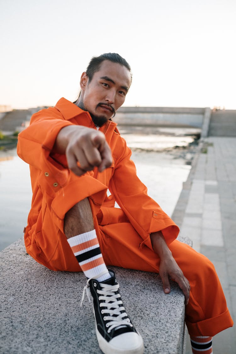 A Man In Orange Jumpsuit Sitting On Concrete Wall Pointing His Finger