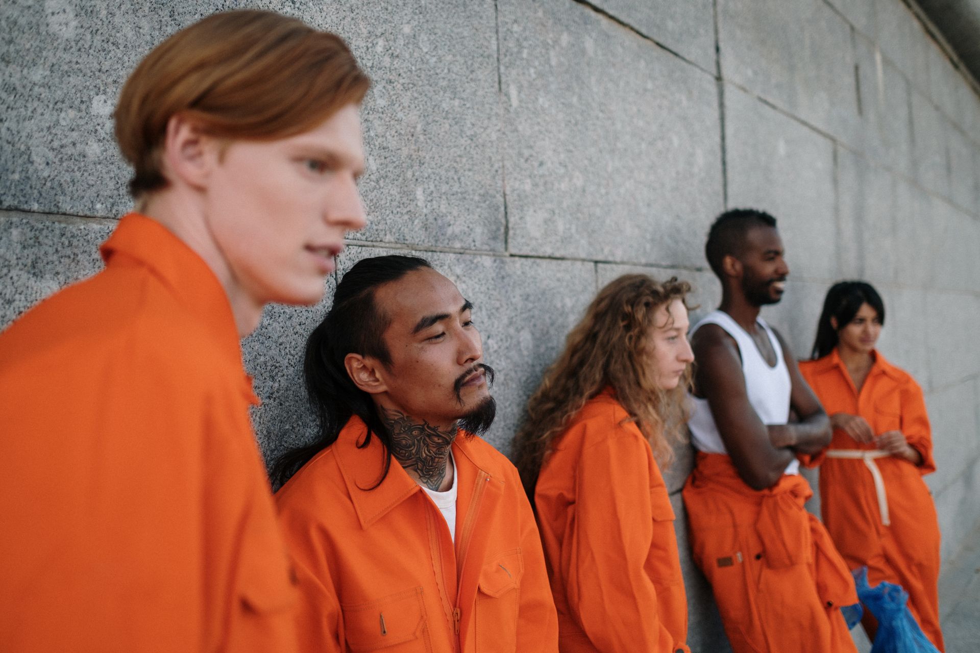 A diverse group of people in orange jumpsuits leaning against a stone wall outdoors.
