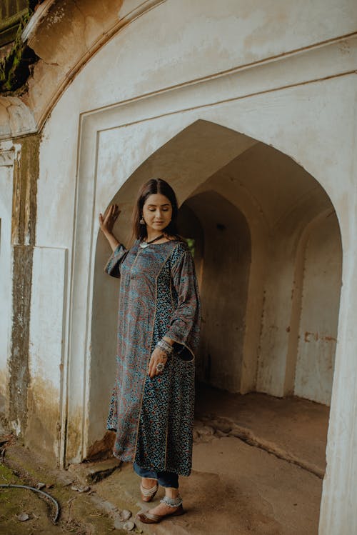Full body of young smiling female in ornamental traditional Muslim apparel with sandals standing in arched vaults of doorway of abandoned building