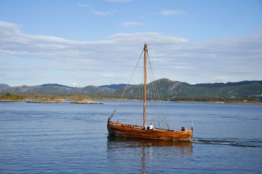 A classic wooden sailboat glides across a tranquil bay with mountainous backdrop under a clear sky.