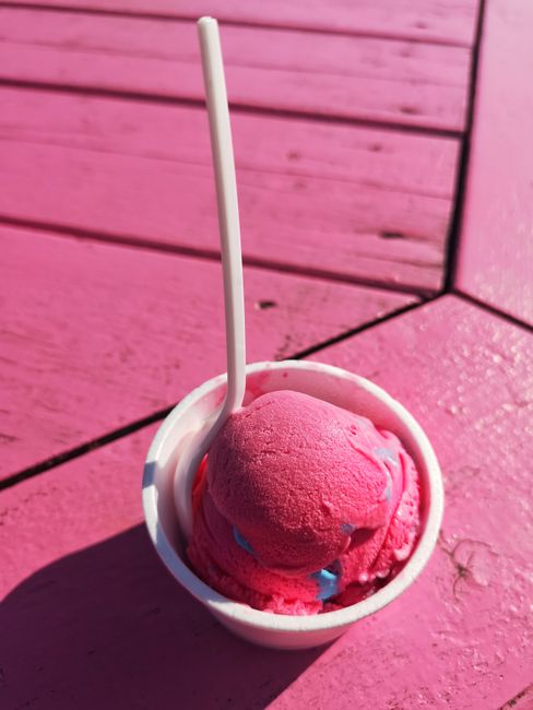 Close-up of a pink ice cream scoop in a cup with a spoon, on a pink wooden table under sunlight.