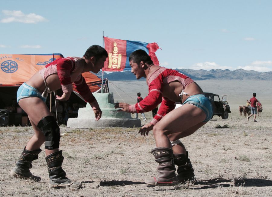 Two men compete in traditional Mongolian wrestling at a festival outdoors with cultural elements visible.