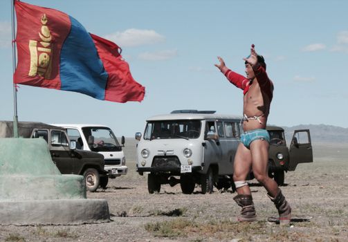 Mongolian wrestler performing outdoors near the national flag with vehicles in the background.