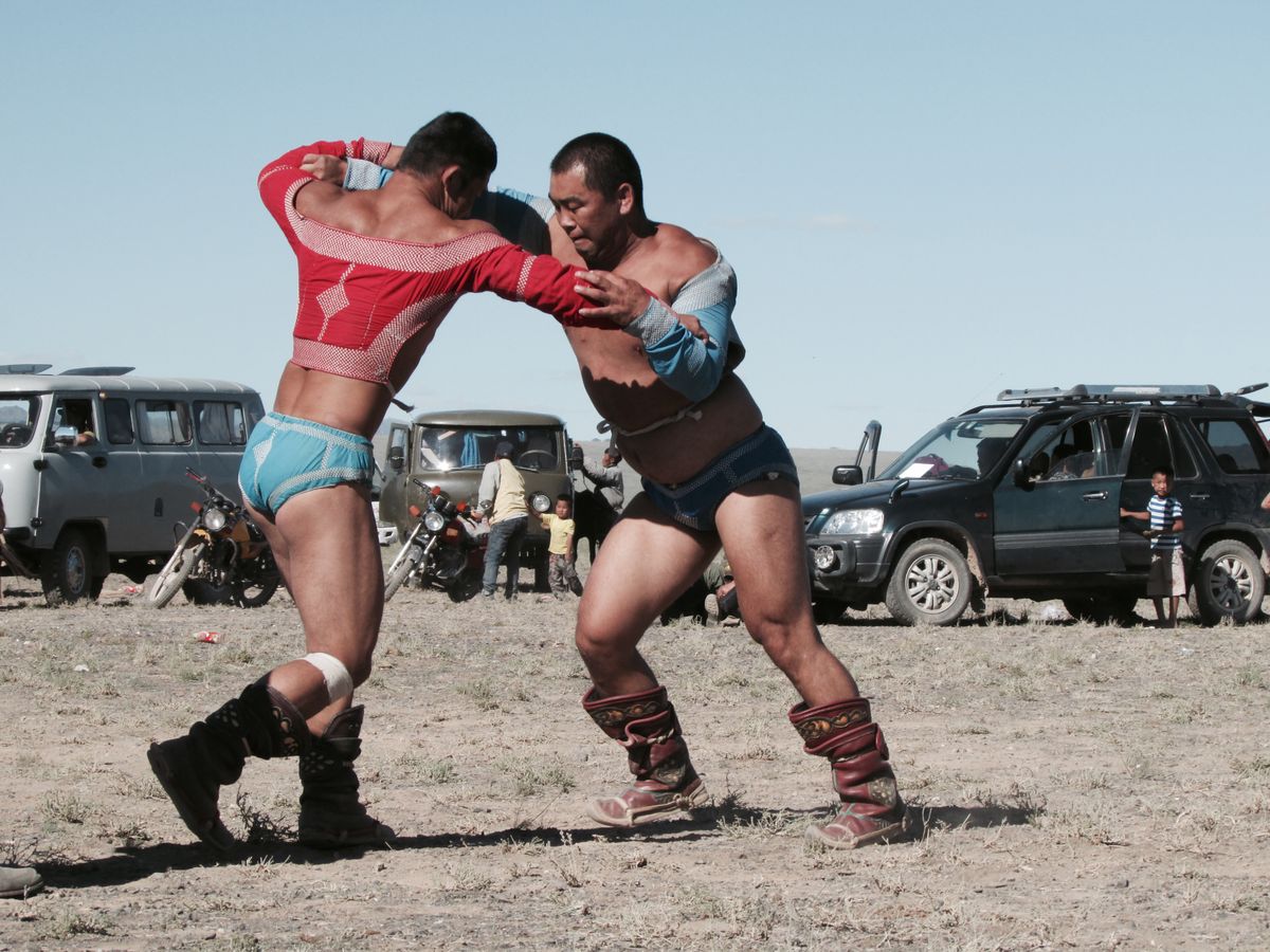 Two Mongolian wrestlers mid-bout, one in a red zodog jacket and one in blue, spectators and vehicles visible behind them