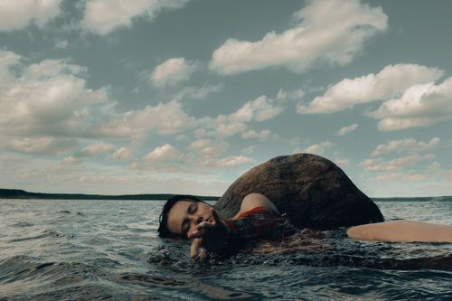 Free A serene scene of a woman resting with eyes closed against a rock in the ocean under a cloudy sky. Stock Photo