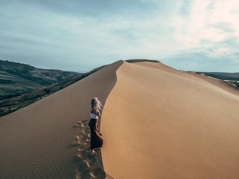 A woman in a white top and long skirt traverses picturesque sand dunes under a cloudy sky. Perfect adventure shot.