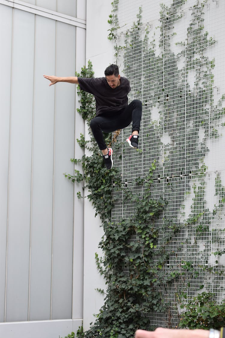 A Man In Black Clothes Jumping Near Climbing Plants On A Wall