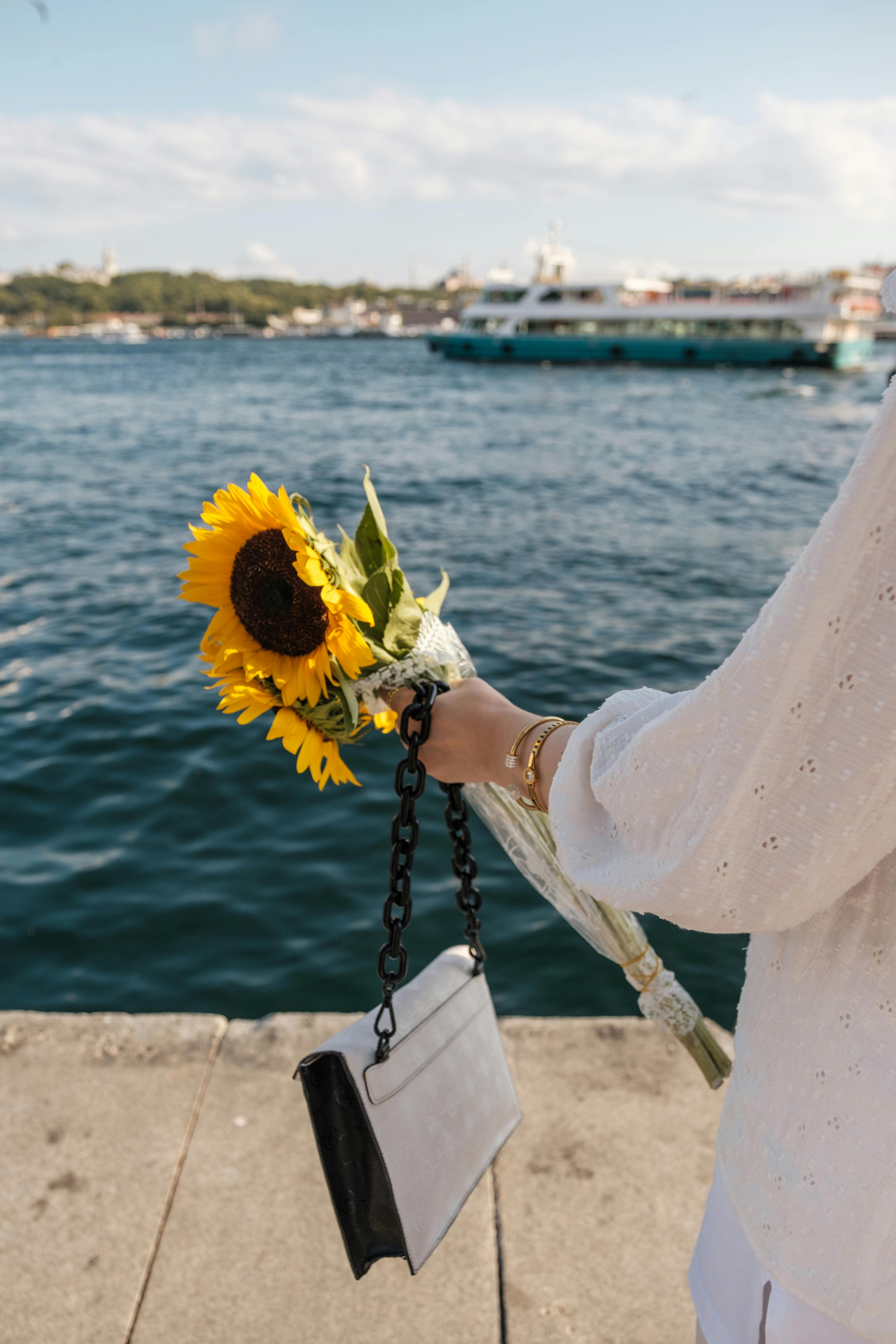 A woman holding sunflowers and a purse by a scenic waterfront with boats.