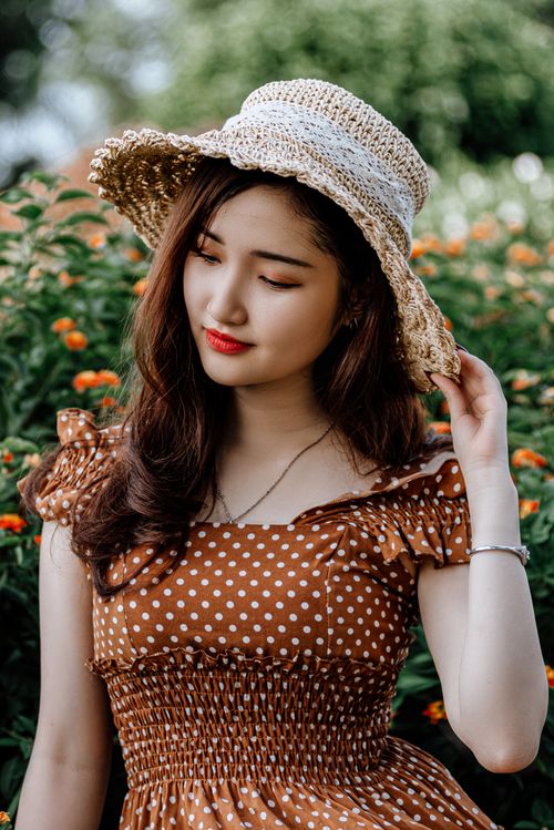Free A young woman in a polka dot dress and sunhat stands amidst vibrant flowers, exuding summertime elegance. Stock Photo