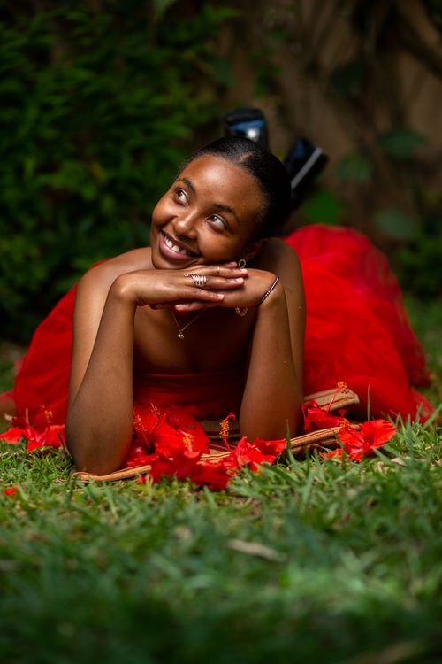 Free A joyful woman lies on grass in a red dress surrounded by vibrant red blossoms, exuding happiness. Stock Photo