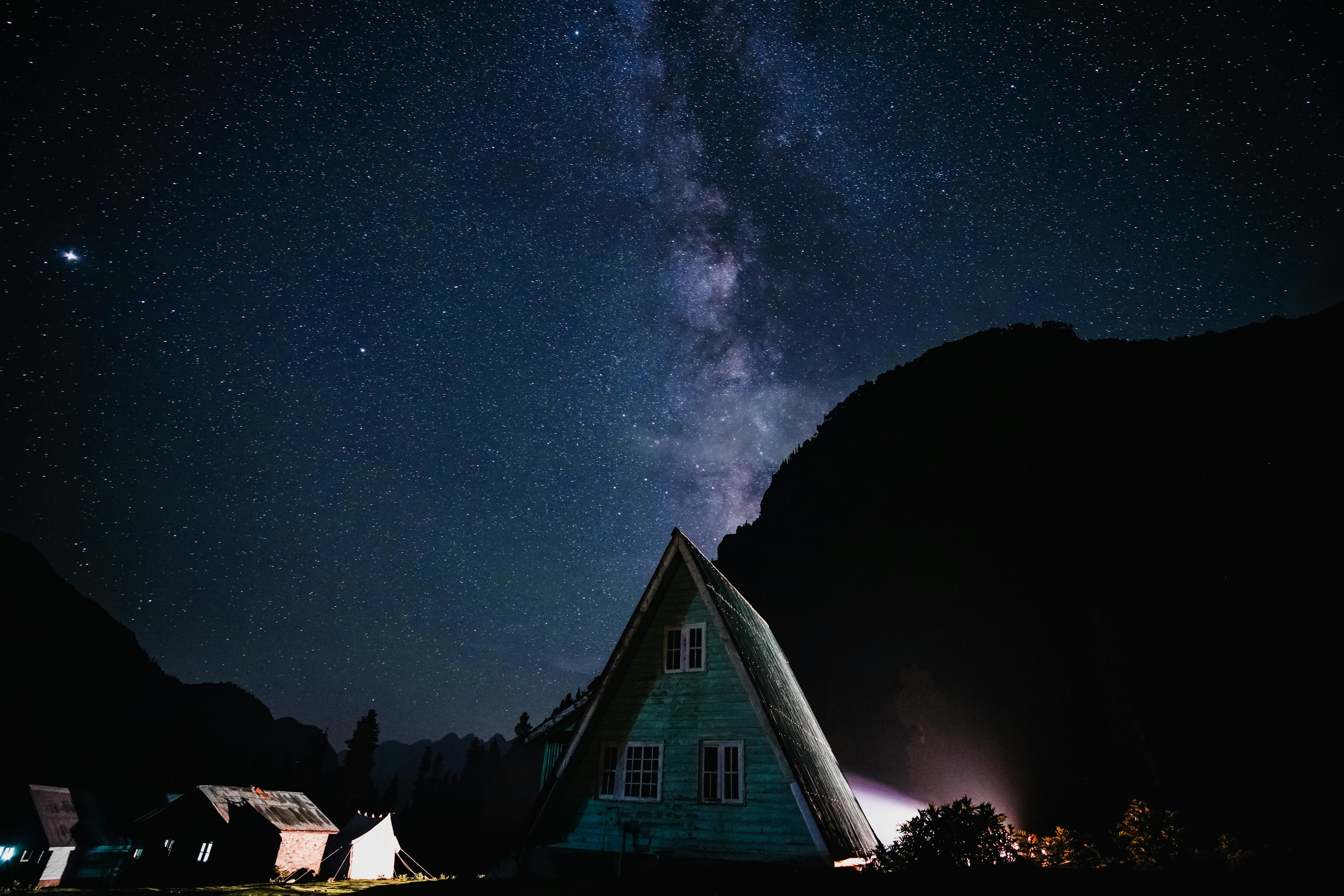 A starry night sky with the Milky Way over an A-frame cabin in a quiet mountain setting.