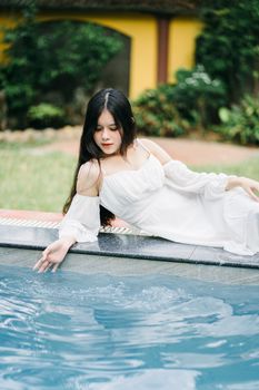 A beautiful woman in a white dress relaxing by the poolside, evoking a sense of elegance and tranquility.