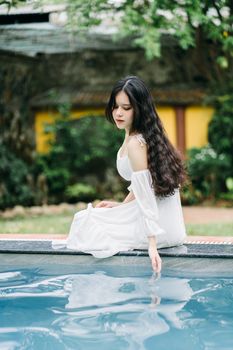 Charming Asian female with black long hair in white dress sitting on poolside near building and trees on summer day