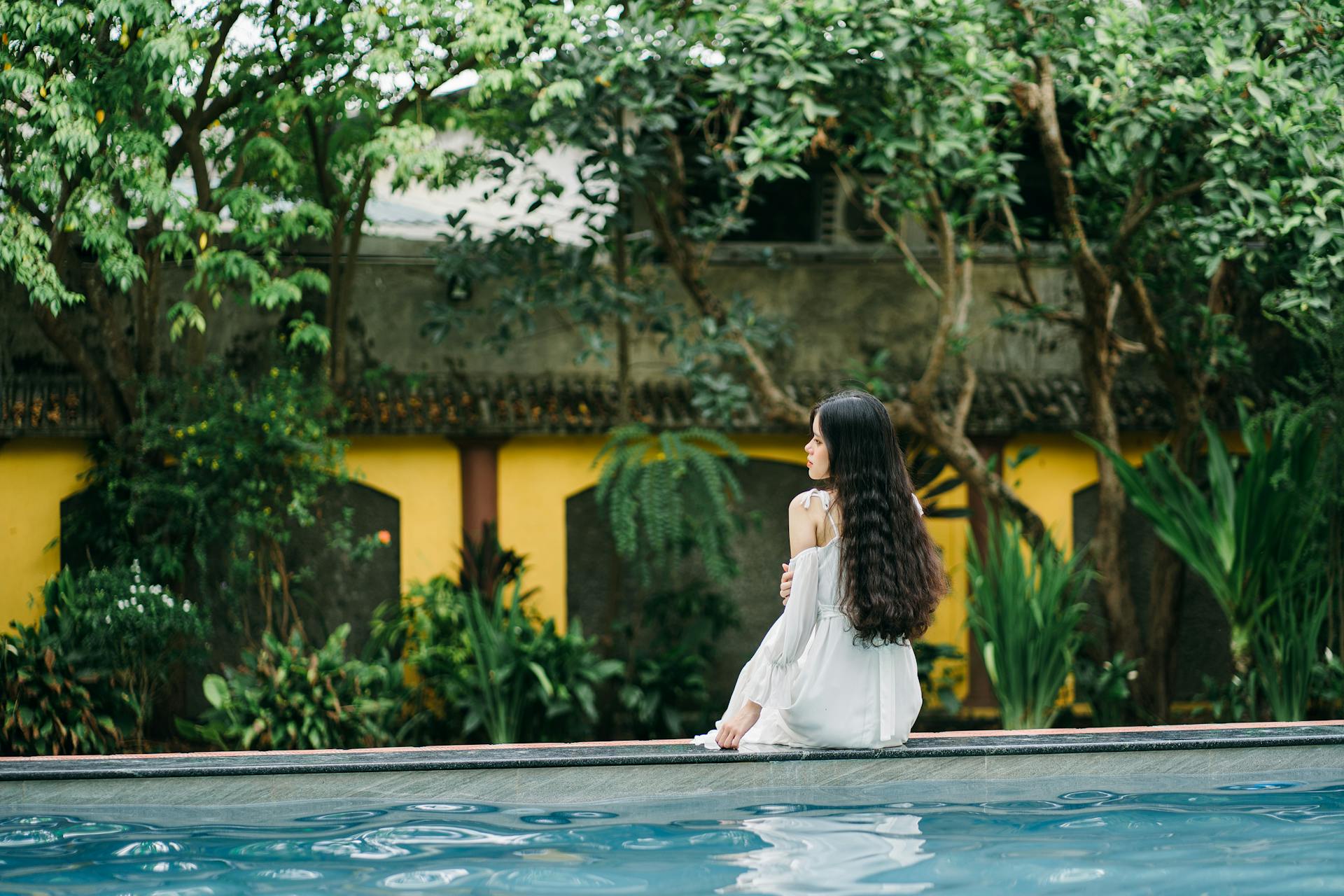 Back view of unrecognizable female with black hair sitting on edge of swimming pool on terrace with building and trees