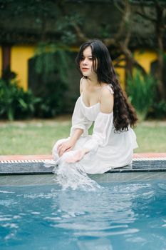 Attractive Asian female in white dress sitting on poolside while touching water on terrace with building and trees on summer day