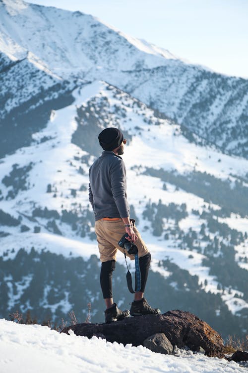 Pria Mengenakan Kemeja Lengan Panjang Abu Abu Dan Celana Pendek Coklat Memegang Kamera Dslr Hitam Di Gunung