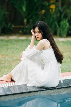 Full body side view of barefoot Asian female in white dress sitting on edge of poolside on terrace with green trees