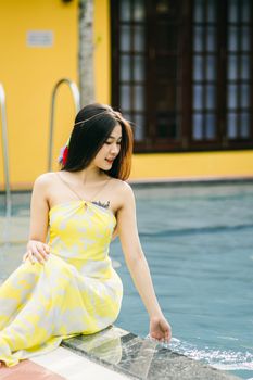 Attractive Asian female in sundress splashing water while sitting on edge of swimming pool on terrace with building on summer day