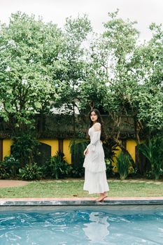 Full body of barefoot female in long white dress standing on edge of swimming pool on terrace with building on summer day