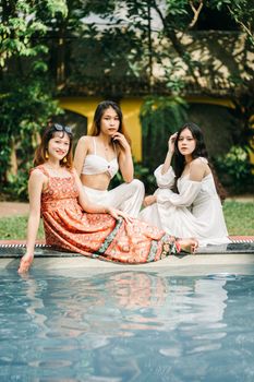 Group of Asian female friends in sundresses looking at camera while sitting together on poolside on terrace with green trees