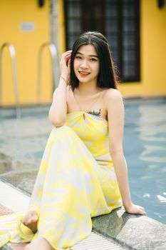 Positive barefoot Asian female in sundress looking at camera with smile while sitting on poolside near building on summer day
