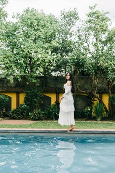 Full body side view of barefoot female in white dress standing on edge of swimming pool near building on summer day
