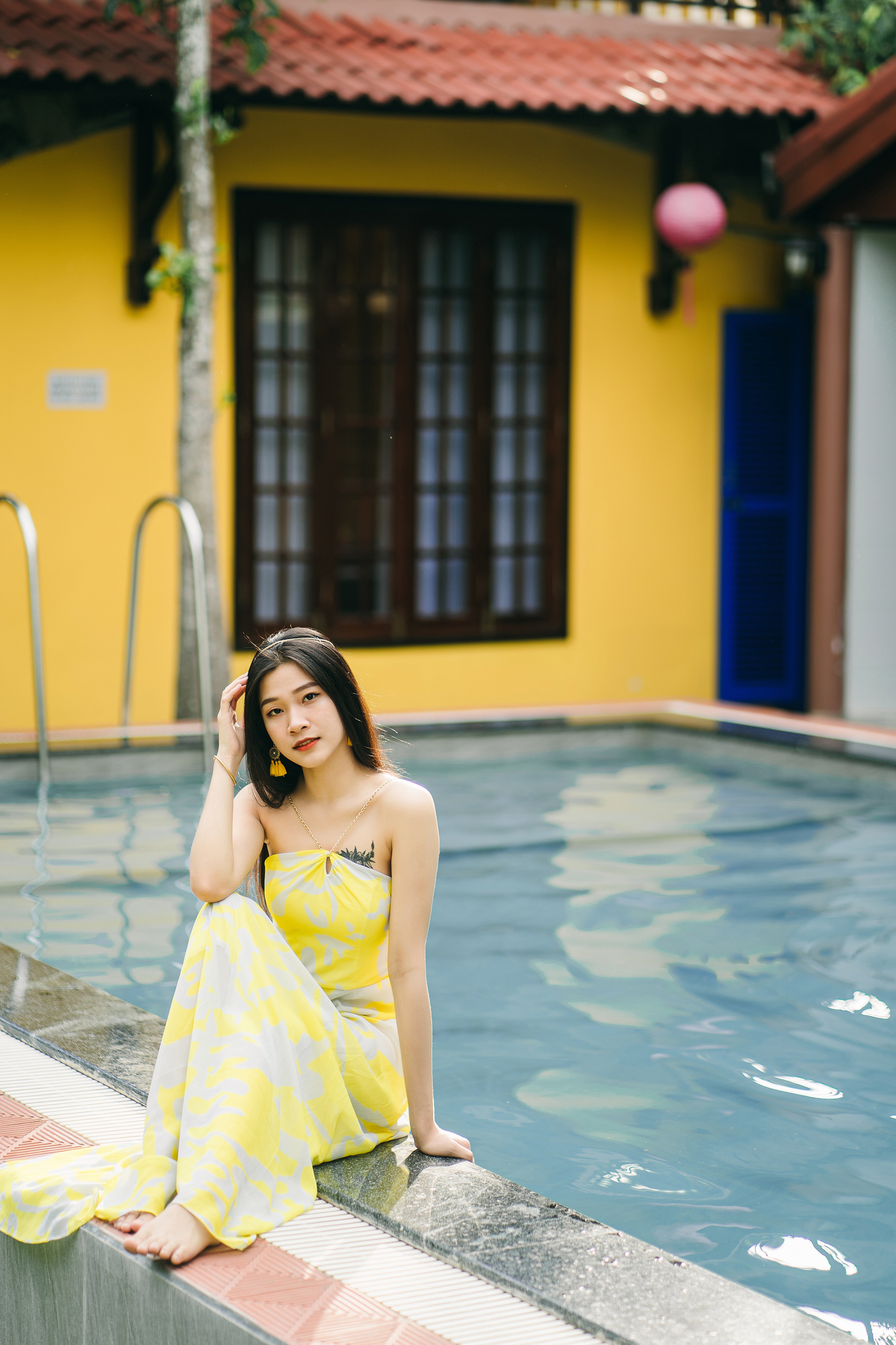Full body of barefoot Asian female in yellow sundress looking at camera while sitting on poolside near building on summer day