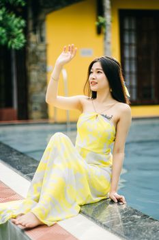 Full body of barefoot Asian female in yellow sundress sitting on edge of swimming pool with raised arm on terrace with building