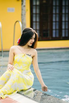 Positive Asian female with dark hair in yellow sundress sitting on edge of swimming pool near building on summer day