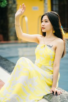 Feminine Asian female with dark hair wearing sundress sitting on poolside with raised arm on terrace with building on summer day