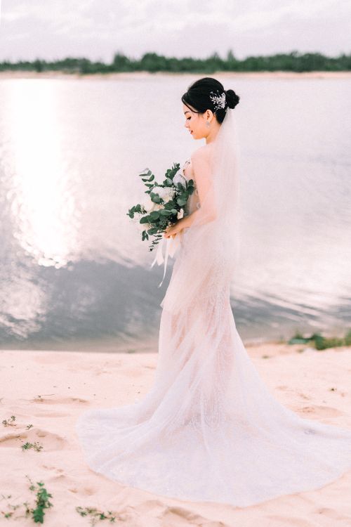 Free A bride in a beautiful wedding dress holding a bouquet, standing by a serene lake at sunset. Stock Photo